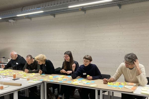 Sechs Personen sitzen an einem langen Tisch und schreiben und kleben Post-Its auf ein Plakat