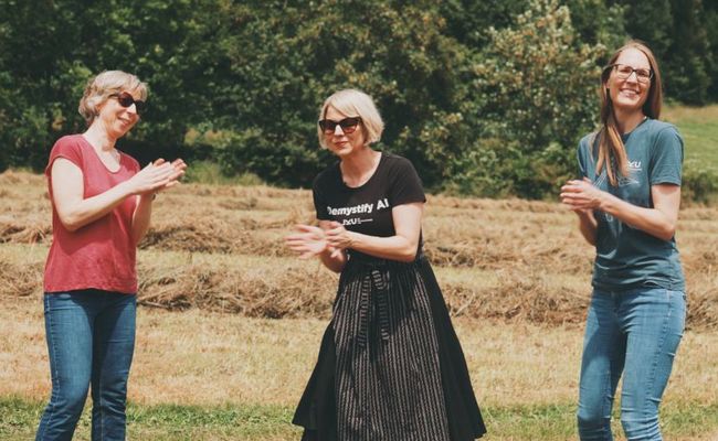 Drei Frauen stehen tanzend und klatschend auf einem frisch geheuten Feld