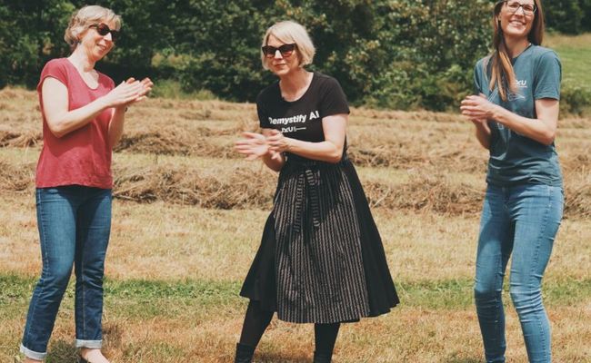 Drei Frauen stehen tanzend und klatschend auf einem frisch geheuten Feld