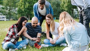 lachende Studierendengruppe, die auf Wiese sitzt und gemeinsam auf ein Handy schaut