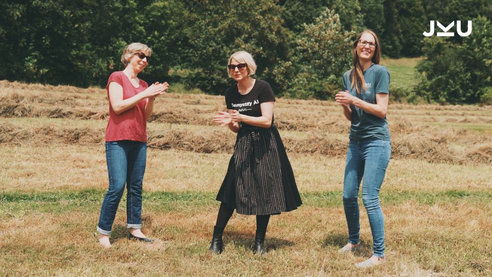 Drei Frauen stehen tanzend und klatschend auf einem frisch geheuten Feld