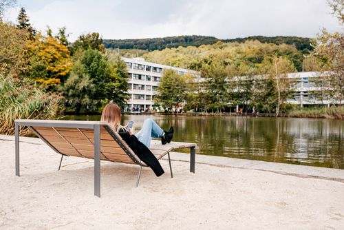 Eine Studentin sitzt auf eine Liegebank und blickt Richtung Uni-Teich