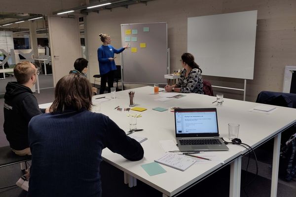 Frau steht vor grauer, mit Post-Its beklebter Wand, stellt diese vier am Tisch sitzenden Personen vor 